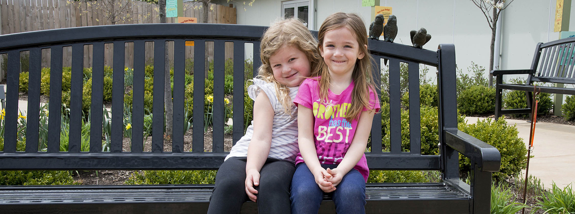 Two young girls sit next to eat other, smiling on a bench. 