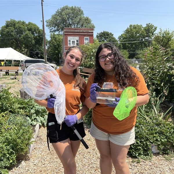 Two high school-aged students stand in a garden with butterfly nets. 