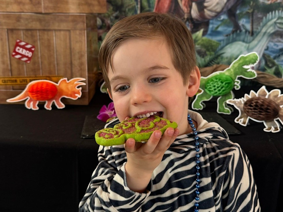 A child eats a dinosaur-themed cookie while standing in front of dinosaur decor. 