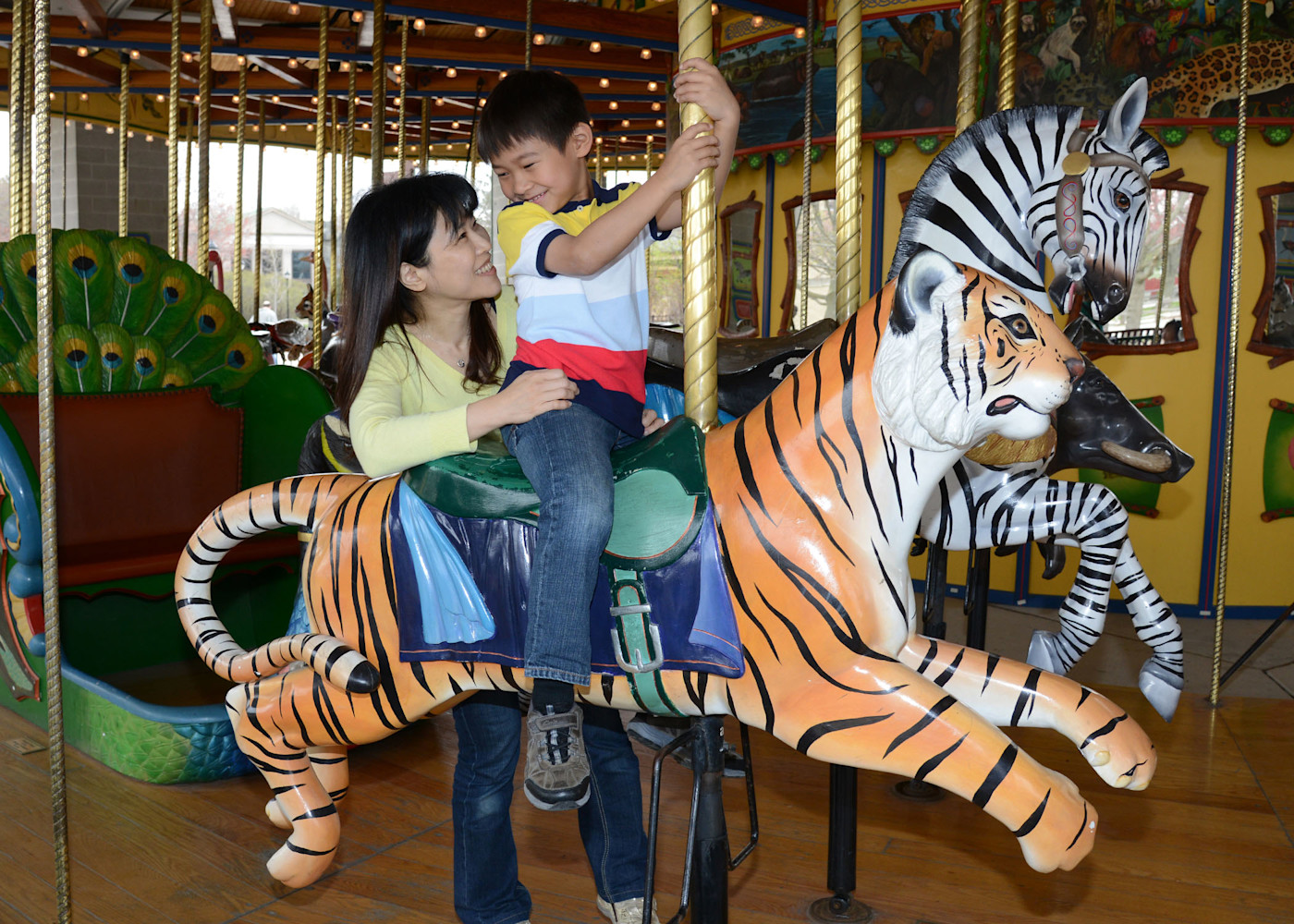 A child sits on the tiger seat of the carousel, their parent standing behind them.