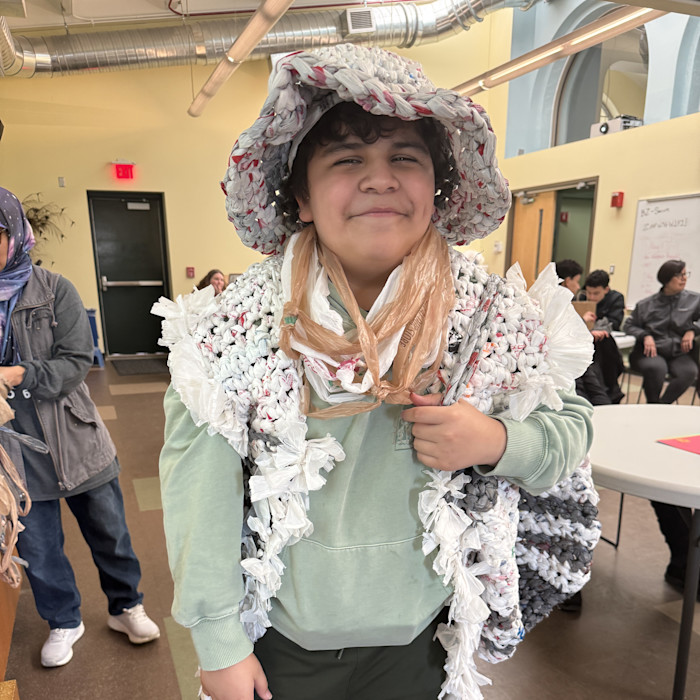 A middle-school aged student wears a vest and hat made out of recycled materials. 
