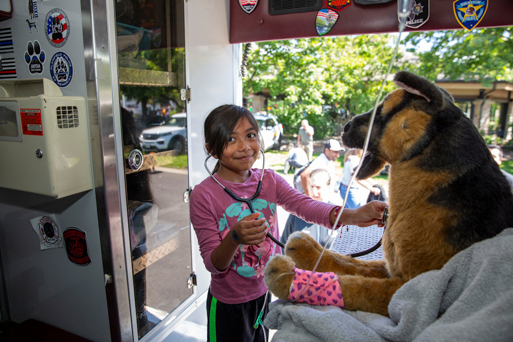 A child wearing a stethoscope stands in an ambulance, pretending to help a stuffed animal. 